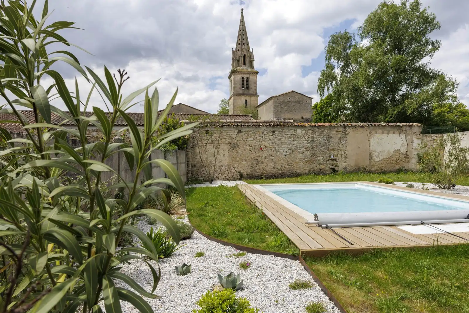 Garden with a pool, a wooden deck and gravel beds, with a church spire behind